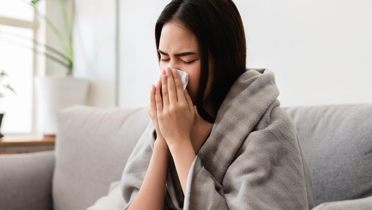 Photo of woman, sitting on couch with a blanket wrapped around her shoulders, blowing her nose in to a tissue. 