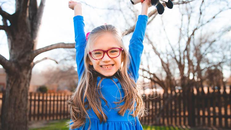 Photo of the 2023 CMN Champion Child Ella with her arms raised triumphantly. Standing in her backyard holding a stuff panda with a pink shirt in her left hand. Clear sky, green grass, wooden mid-height picket fence and tree are visible in the background. Sun is directly behind Ella. 