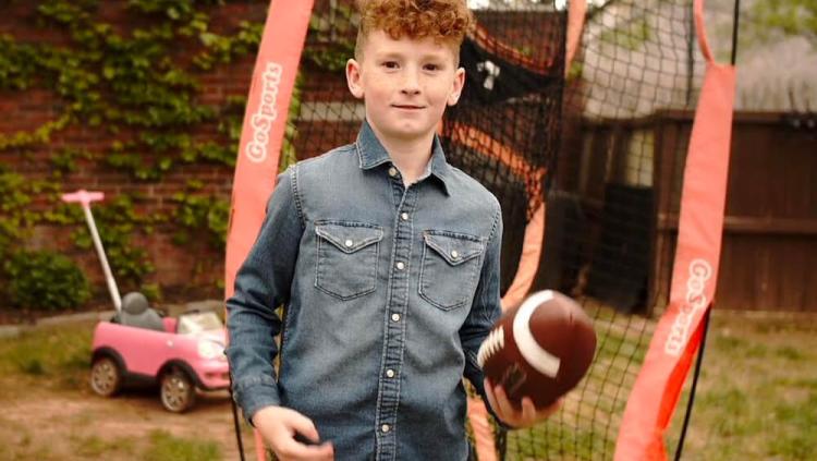 Photo of the 2022 CMN Champion Child Finn in his backyard holding a football in his left hand. Visible in the background are a football passing practice net, a pink child's ride-along care with t-handle, ivy on a brick wall and privacy fencing. 